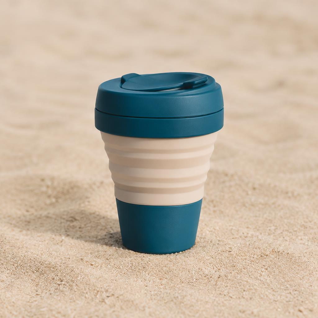Collapsible travel cup in ocean blue and sand beige with a lid, standing on sand